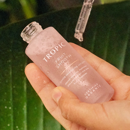 Hand holding a Tropic Fruit Smooth gentle resurfacing serum bottle with water droplets, with a glass dropper above and a green leaf background.